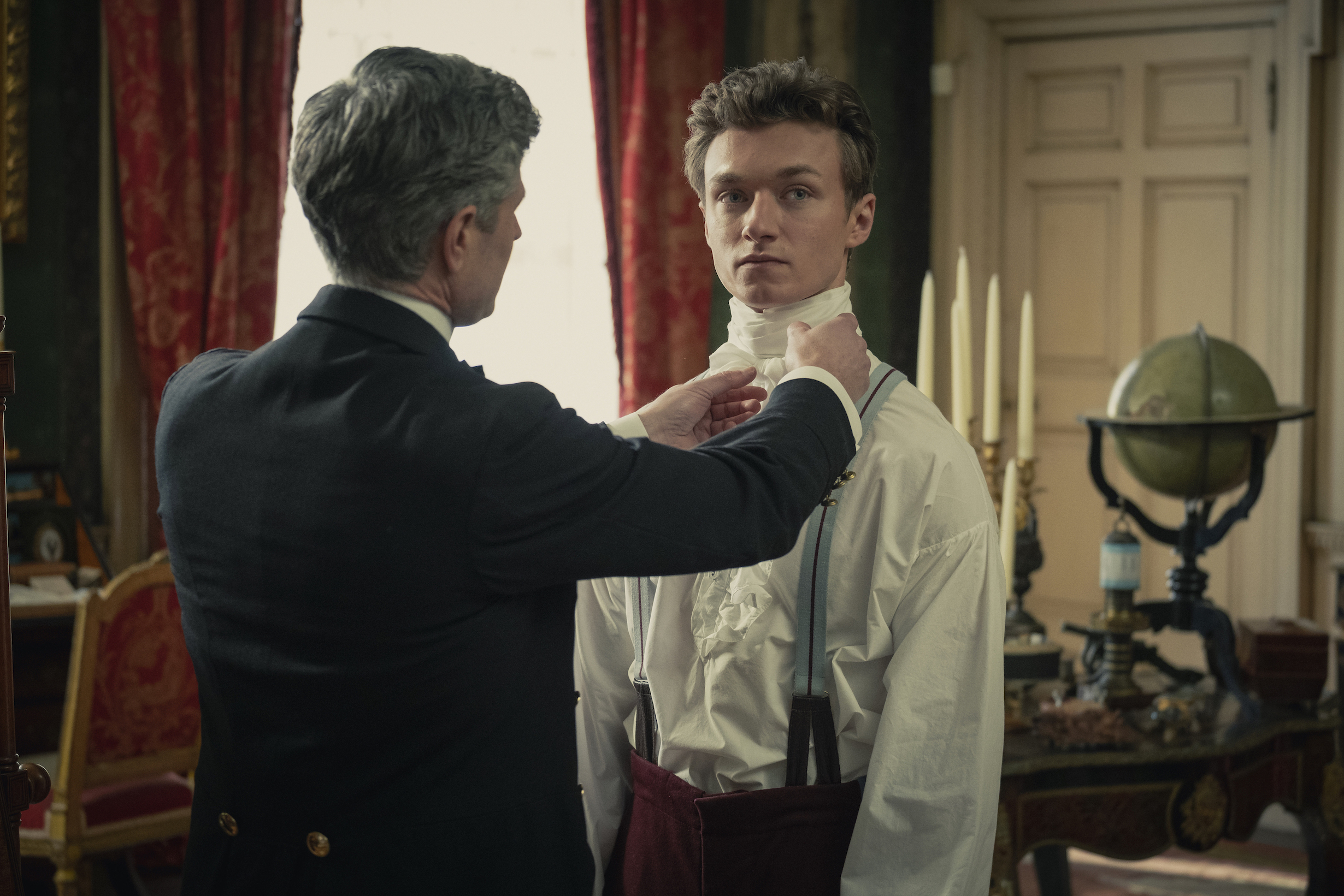 A man being helped to dress in a ruffled shirt in an opulent room.