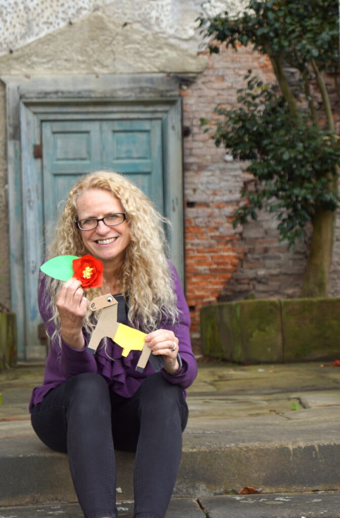 Dr Suzanne Carter sits on a step in front of an old door holding a paper camellia and horse.